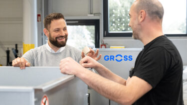 Ein GROSS Mitarbeiter berät neben einer Brikettpresse einen Kunden mit einem Brikett in seinen Händen